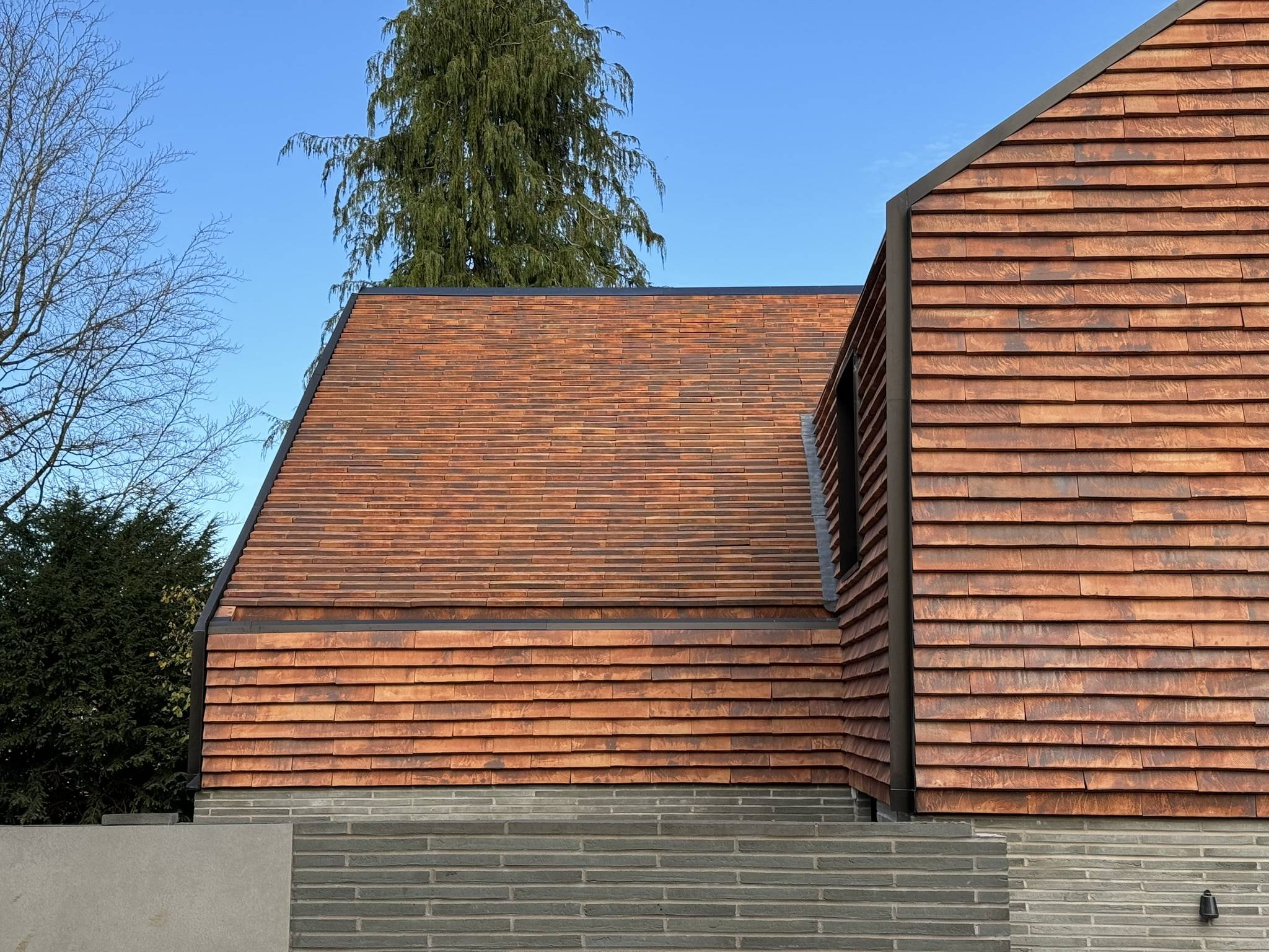 Close-up of a modern building’s sloped and vertical roof sections with red terracotta tiles, grey brick walls, and a clear blue sky, architecture by Gresford Architects.
