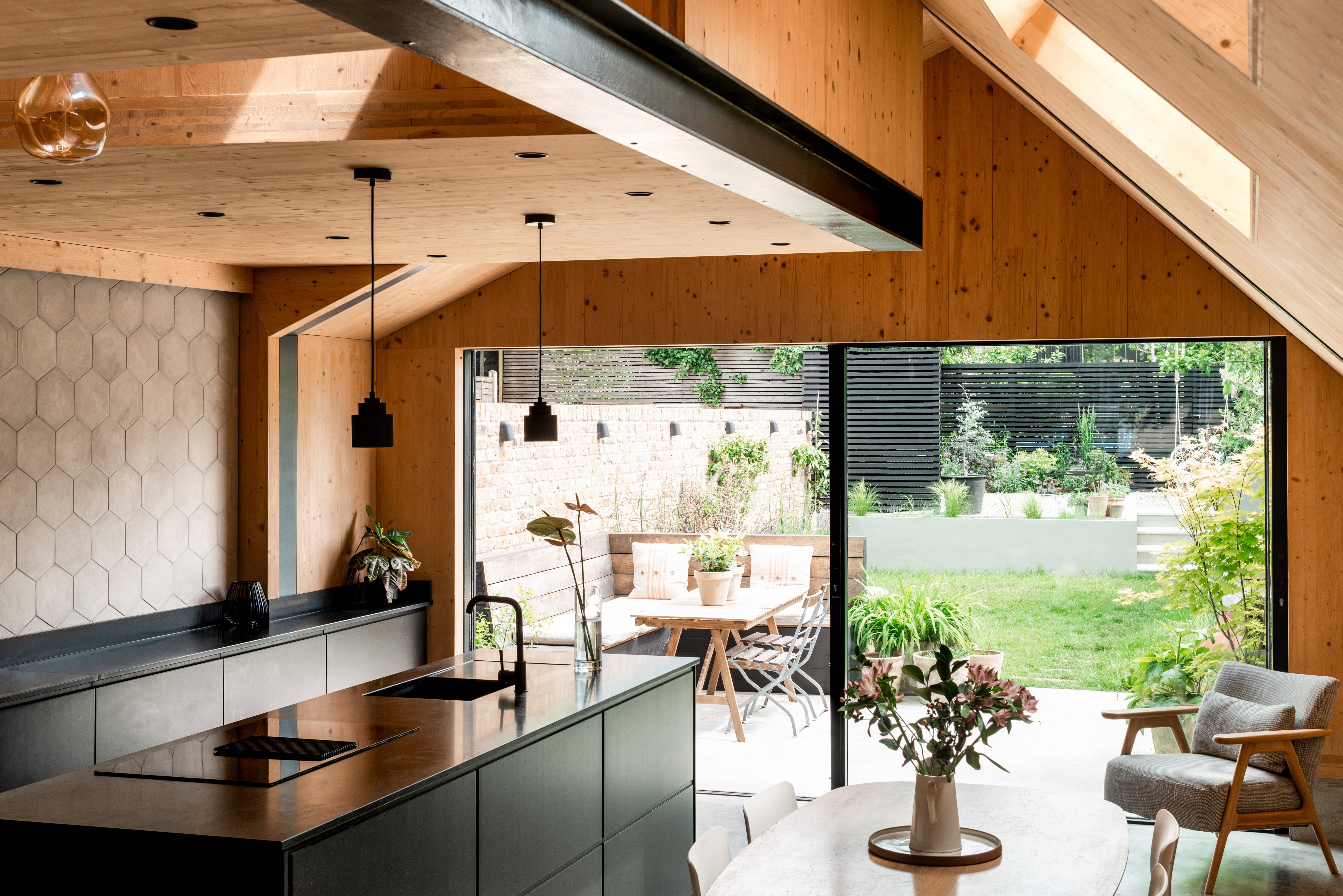Open-plan kitchen with black cabinetry, wooden ceiling and walls, and large sliding glass doors leading to a garden; architecture by Gresford Architects.