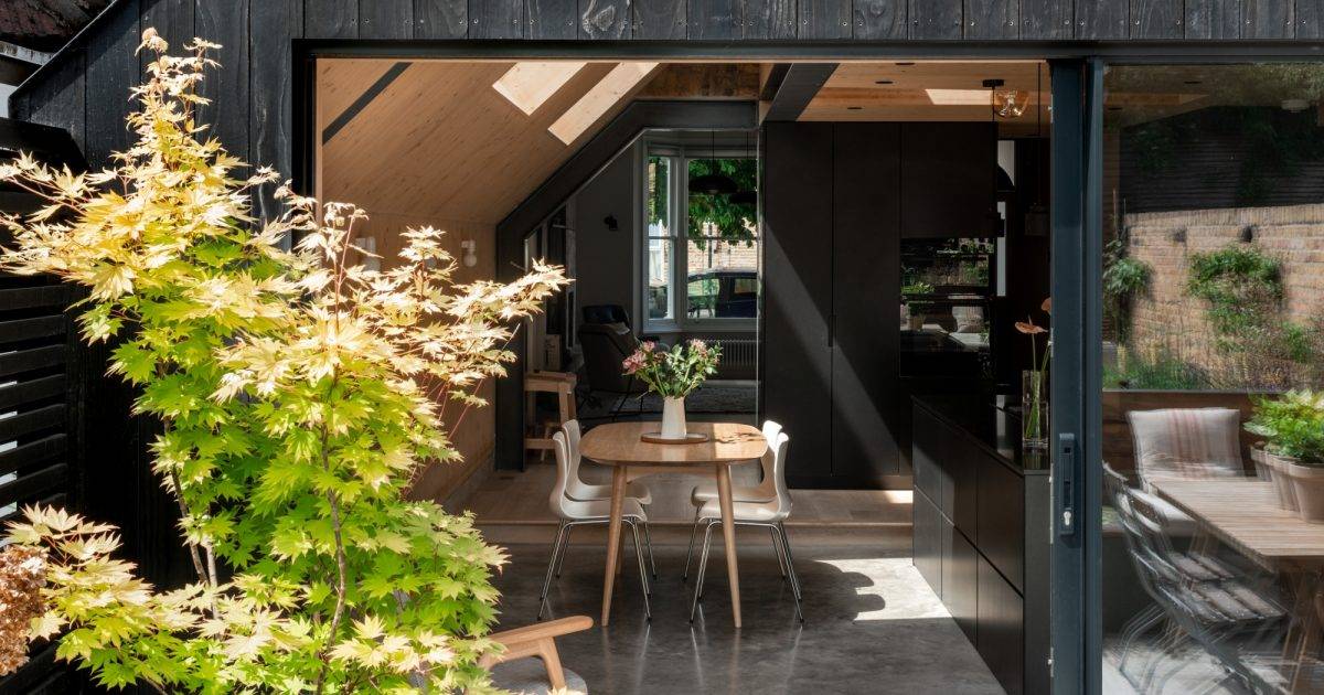 Gresford Architects Ltd — A modern interior with a round wooden dining table and white chairs, black cabinetry, and wooden ceiling, with a view to an outdoor garden; architectu