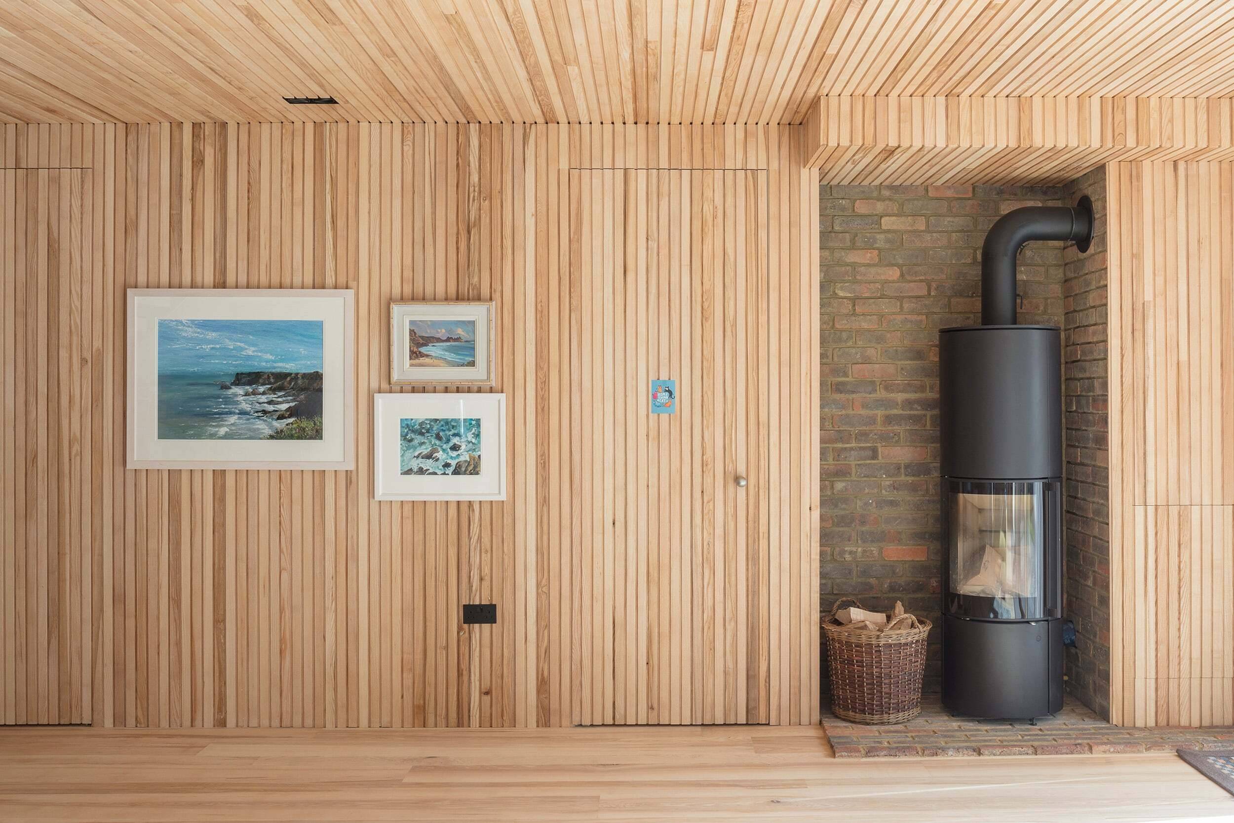 A wood-paneled wall with three framed landscape pictures in a living space, featuring a modern black stove, brick hearth, and a basket, architecture by Gresford Architects.