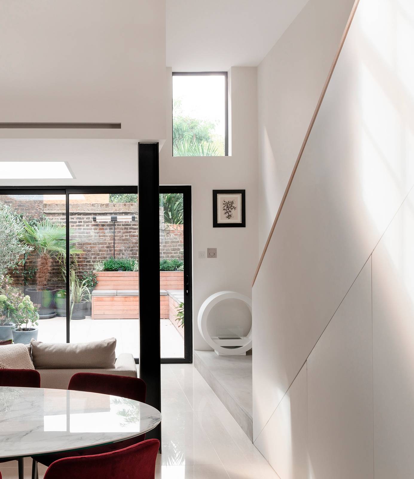 Modern interior with white walls, a large glass door leading to a garden, artwork, and minimalist furniture, architecture by Gresford Architects.
