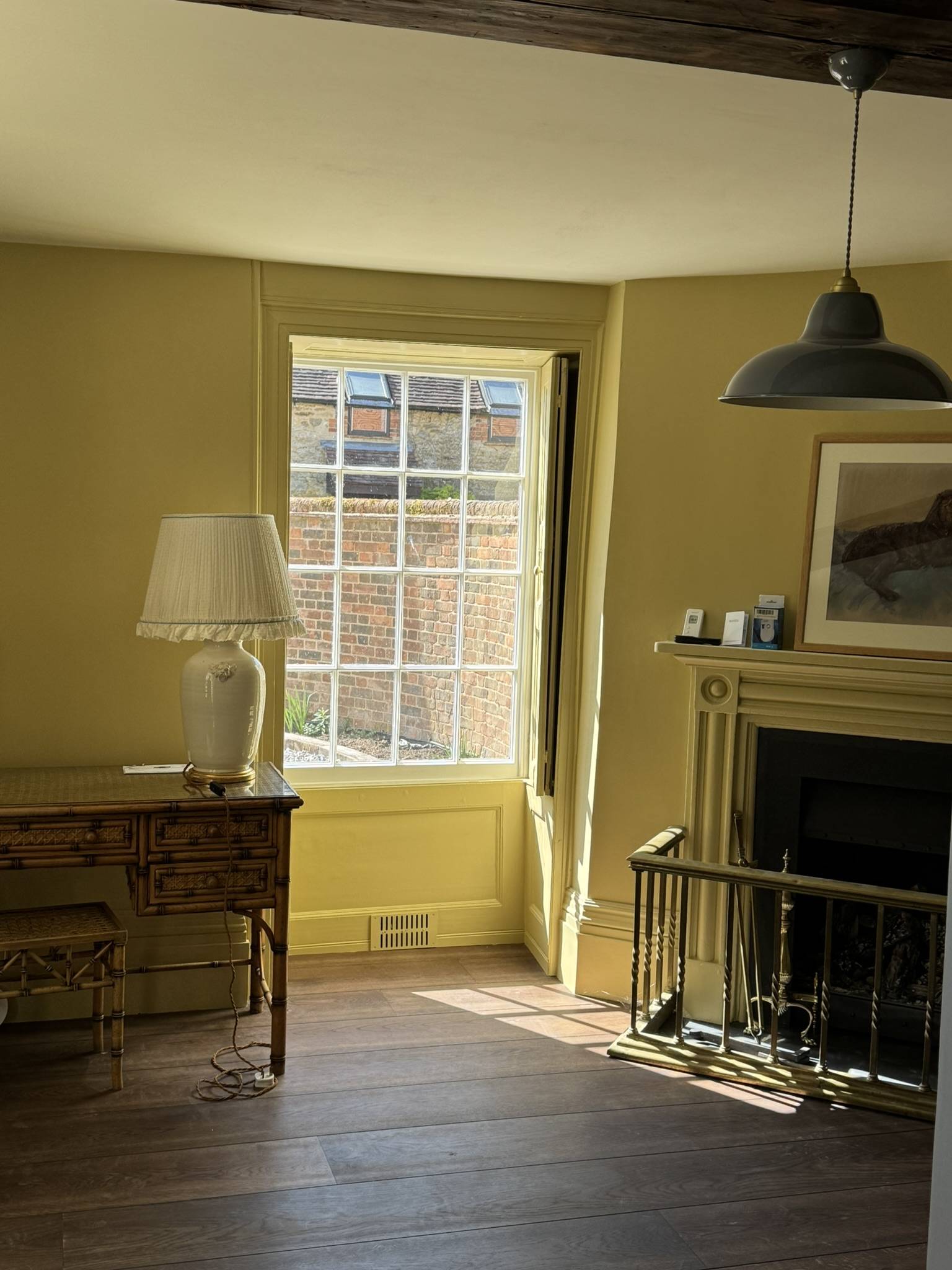 Bright interior with a large window allowing sunlight, a decorative fireplace, a table with a lamp, and wooden flooring; architecture by Gresford Architects.