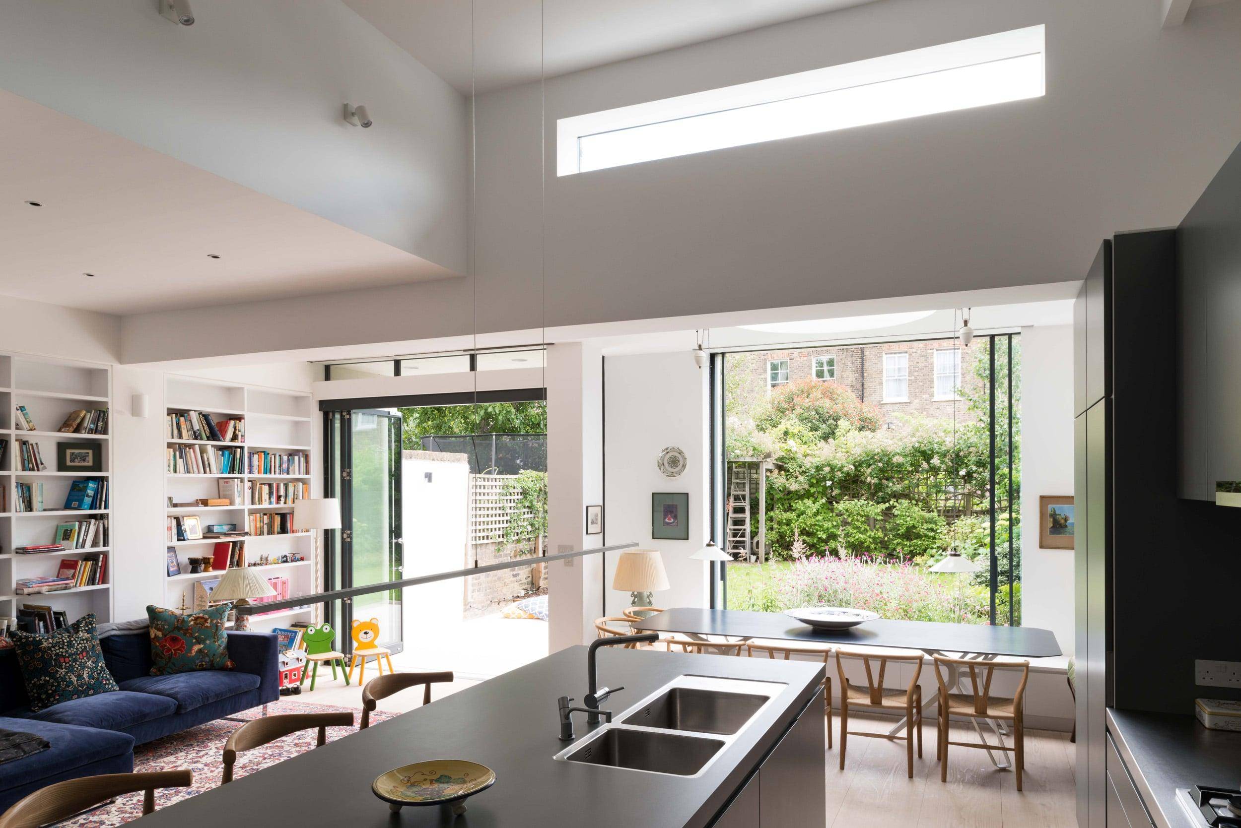 Interior view of a modern, open-plan living space with large windows open to a garden, featuring white walls, built-in bookshelves, and a kitchen island, architecture by Gresford Architects.