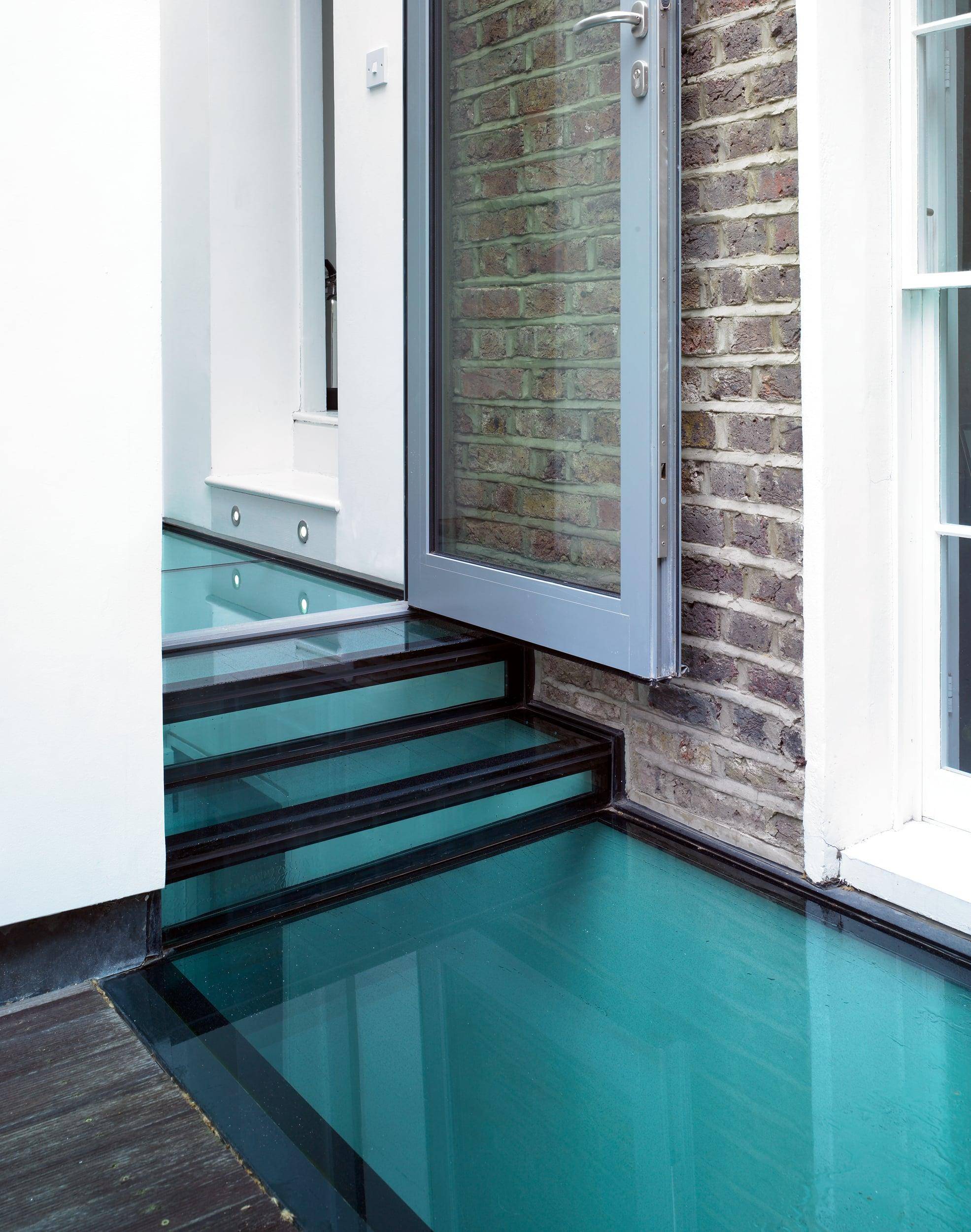 Close-up of a modern glass staircase by Gresford Architects, leading to a brick and white wall, with an open grey door; architecture by Gresford Architects.