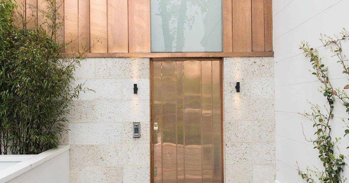 Gresford Architects Ltd — Modern front entrance with copper door, wooden panels, and frosted glass feature, surrounded by greenery; architecture by Gresford Architects.