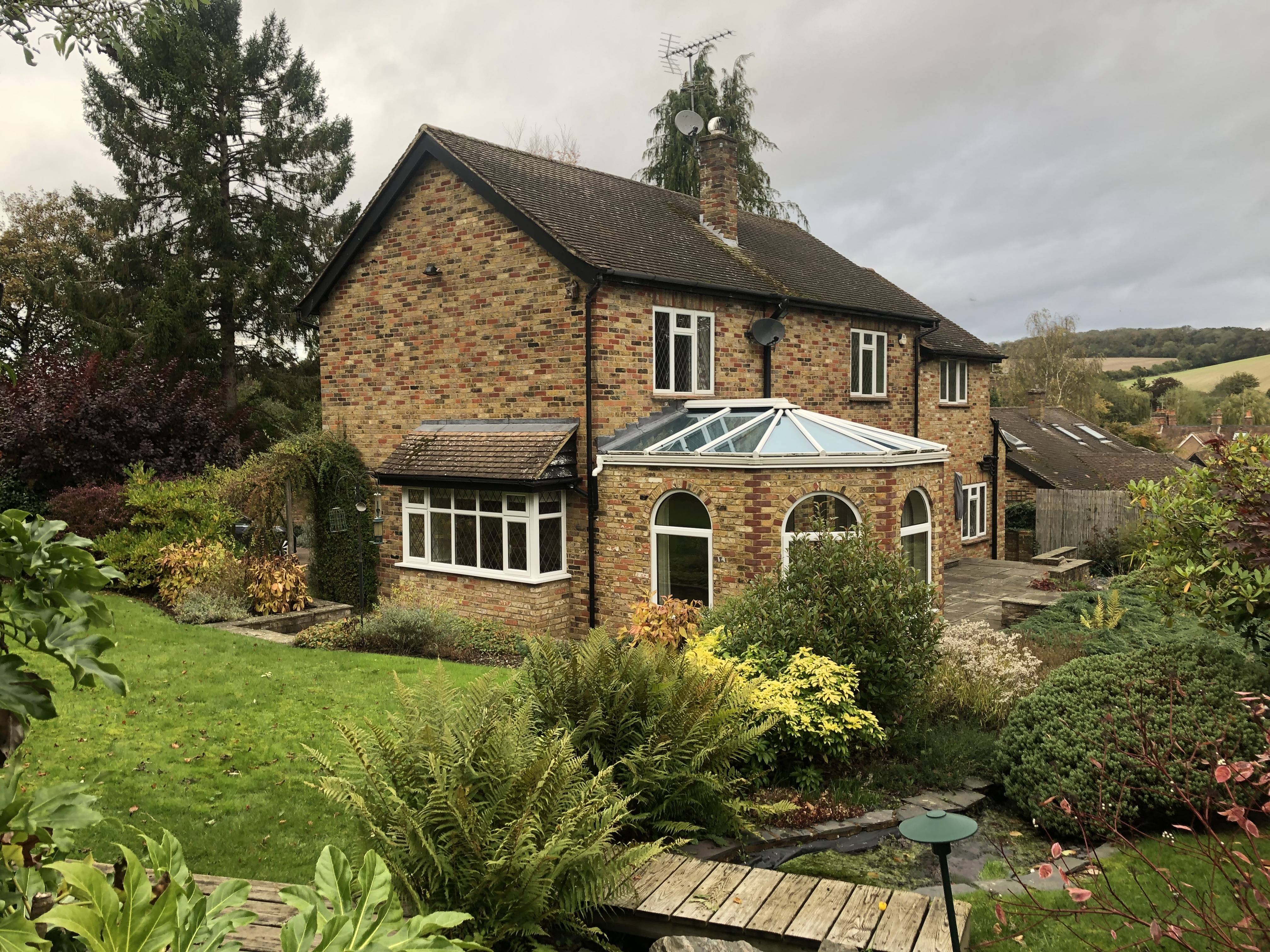 A brick house with a large glass-roofed extension, surrounded by a lush garden with various plants and trees, under a cloudy sky, architecture by Gresford Architects.