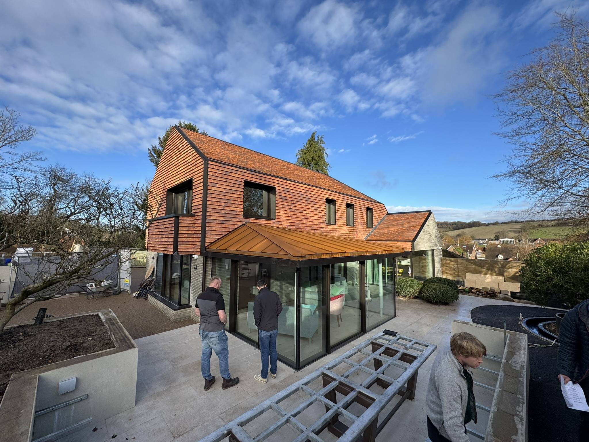 Modern two-story house with large glass windows and a sloped wooden roof; three people stand on a patio, and there’s a partially constructed structure in the foreground. Trees and countryside are in the background under a blue sky.