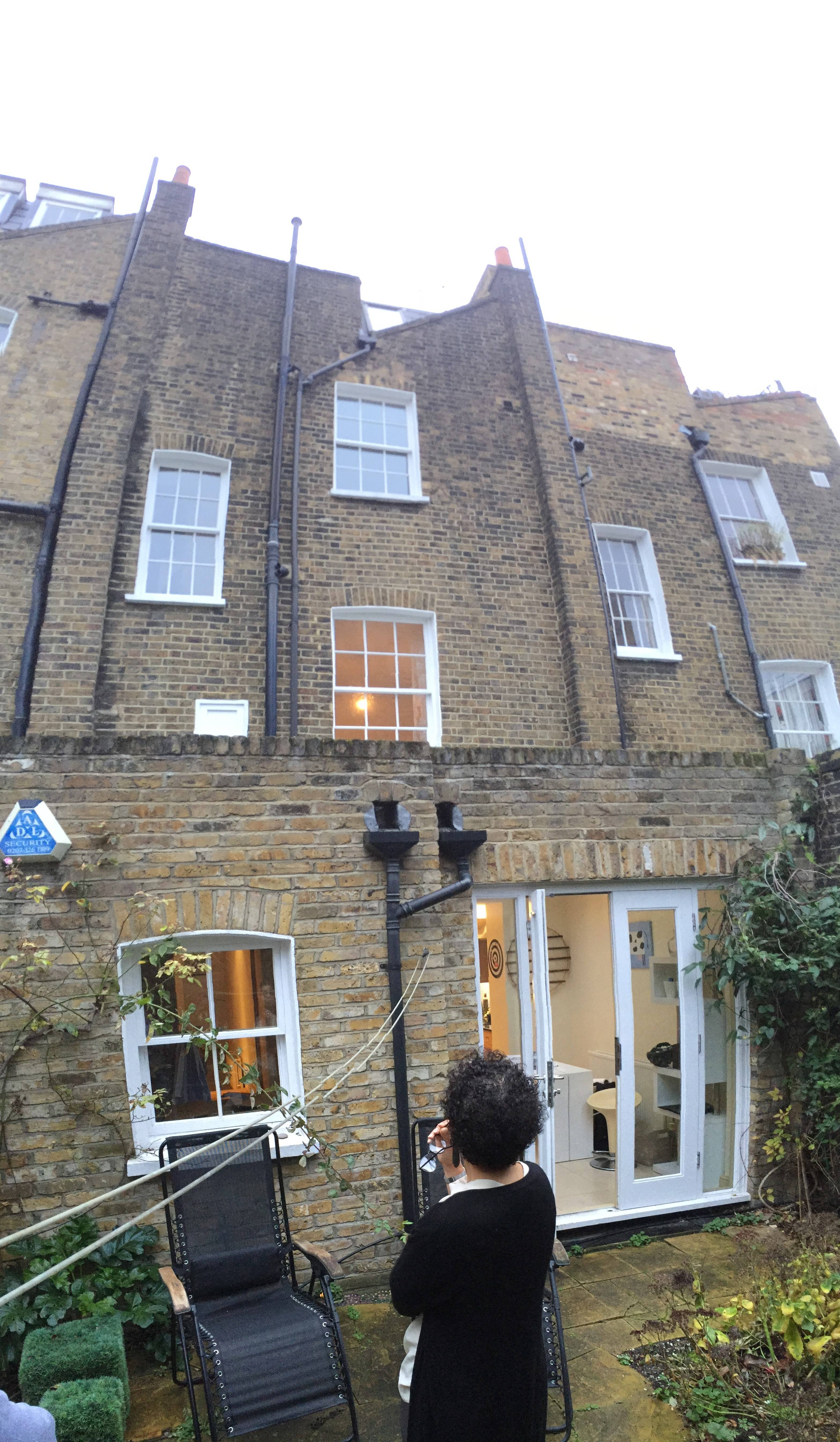 A woman in a black coat stands in a small garden, looking at her phone, with a brick building and a glass extension behind her; architecture by Gresford Architects.