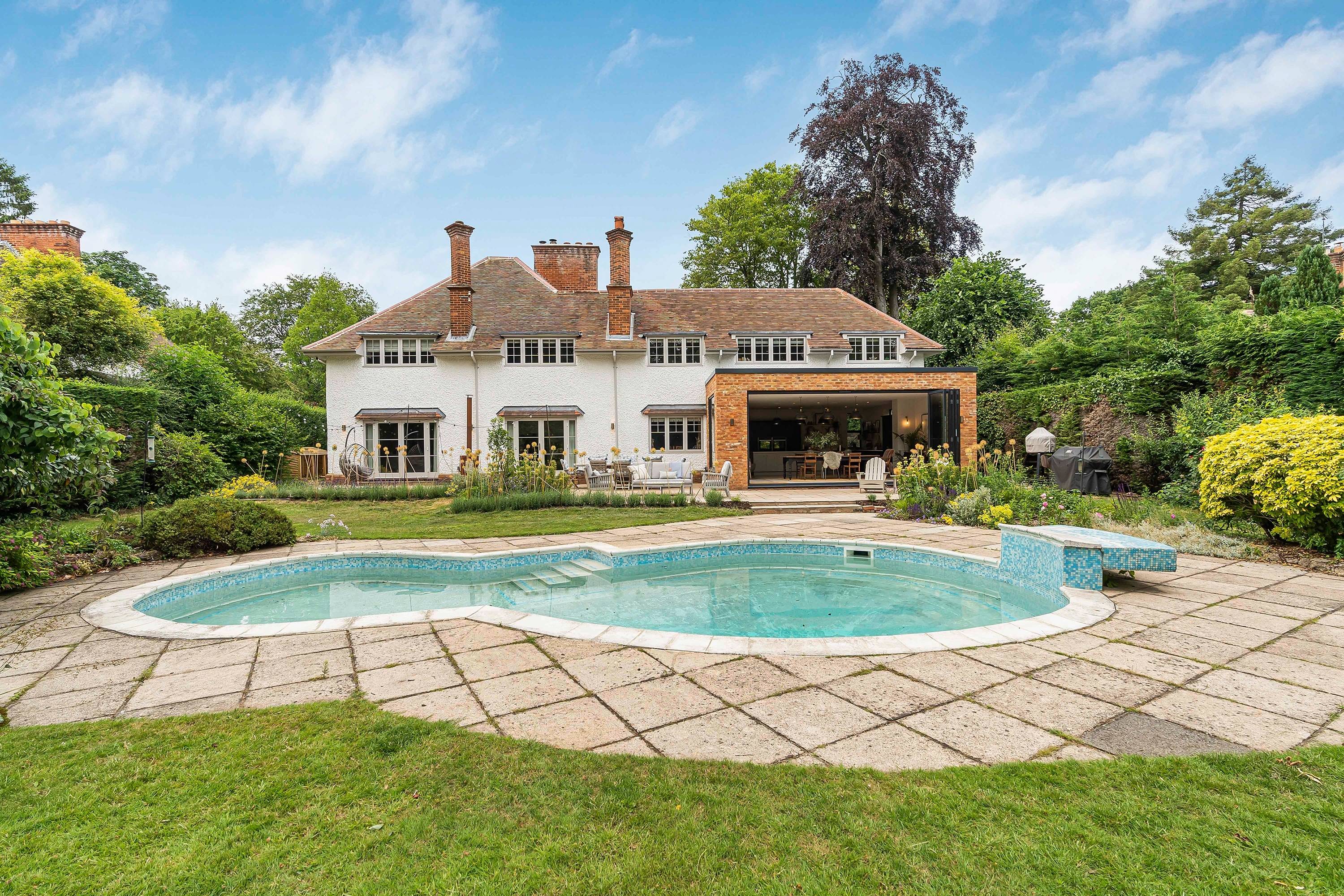 A rear view of a traditional two-storey house with a white facade, multiple chimneys, garden, patio with outdoor furniture, swimming pool, and lush greenery, architecture by Gresford Architects.